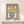 May include: Framed vintage-style poster with purple border featuring yellow flowers with red centers in a blue polka-dotted vase, displayed on light wood console table next to small wrapped gift with greenery, against cream wall with natural lighting