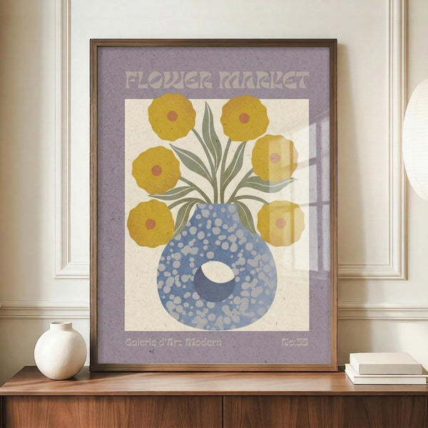 May include: Framed vintage-style poster with purple border featuring yellow flowers in blue polka-dotted vase, displayed on wooden credenza beside white ceramic vase and stacked books against cream wall with white wainscoting in modern living room setting.