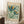 May include: Vintage-style poster with colorful stylized flowers in blues, greens, and orange against cream background, displayed in light wood frame leaning against white wall corner. Small white teacup on wooden block and beige throw blanket visible nearby on white textured rug.