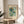 May include: Framed botanical poster with stylized flowers in teal, green, and orange on pale background, displayed on round wooden side table next to beige upholstered chair. Natural sunlight creates shadows across white wall behind artwork. Plant leaves visible in corner of bright, minimalist living space.