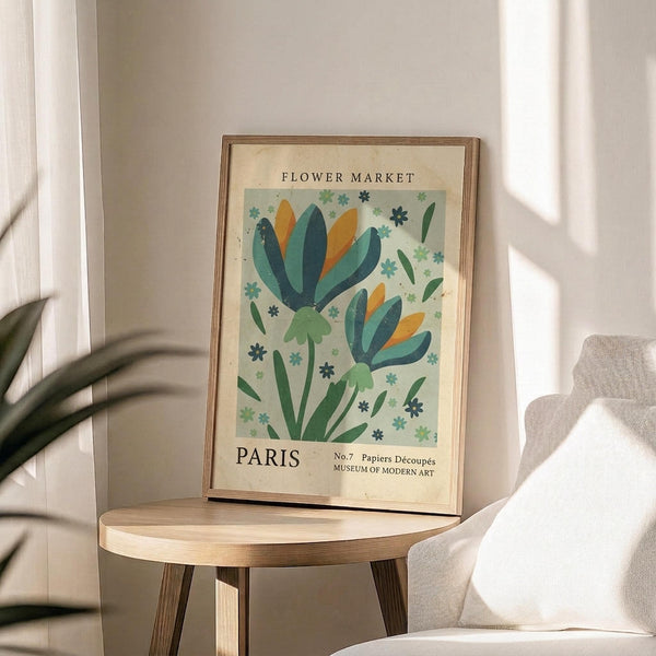 May include: Framed botanical poster with stylized flowers in teal, green, and orange on pale background, displayed on round wooden side table next to beige upholstered chair. Natural sunlight creates shadows across white wall behind artwork. Plant leaves visible in corner of bright, minimalist living space.