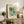 May include: A framed botanical poster with stylized flowers in blue, green, and orange tones displayed on a rustic wooden console table beside a fern plant, books, and ceramic vase in a neutral-toned living room setting with beige furniture and soft natural lighting