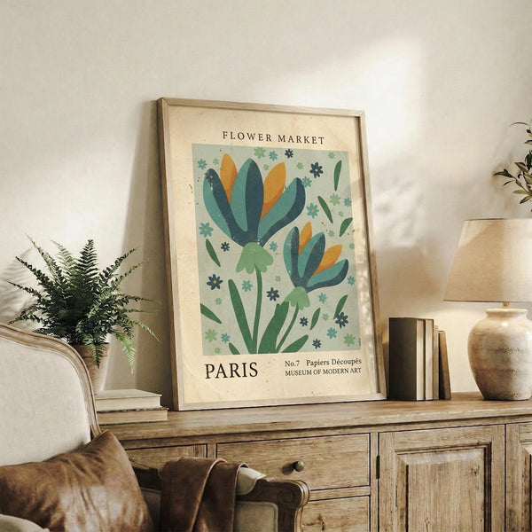 May include: A framed botanical poster with stylized flowers in blue, green, and orange tones displayed on a rustic wooden console table beside a fern plant, books, and ceramic vase in a neutral-toned living room setting with beige furniture and soft natural lighting