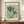 May include: A framed vintage-style botanical poster featuring stylized flowers with teal, orange, and green petals scattered with small decorative blooms, displayed on a wooden sideboard next to a cream ceramic vase and stacked books in a room with white wainscoting and beige walls.