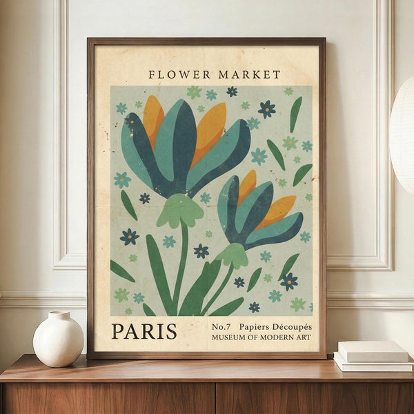 May include: A framed vintage-style botanical poster featuring stylized flowers with teal, orange, and green petals scattered with small decorative blooms, displayed on a wooden sideboard next to a cream ceramic vase and stacked books in a room with white wainscoting and beige walls.