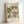 May include: Framed botanical art print leaning against white wall featuring vintage-style floral pattern with green, yellow, and rust-colored flowers and leaves on cream background. Text reads "Flower Market," "Papiers Découpés," "Santorini," "Art Collection," and "Exhibition Flowers" with "No. 18" marking. Light wooden frame with natural wood grain visible.
