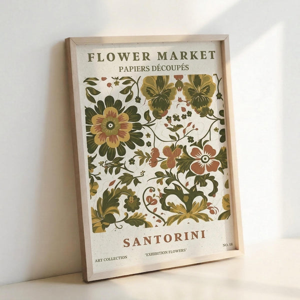 May include: Framed botanical art print leaning against white wall featuring vintage-style floral pattern with green, yellow, and rust-colored flowers and leaves on cream background. Text reads "Flower Market," "Papiers Découpés," "Santorini," "Art Collection," and "Exhibition Flowers" with "No. 18" marking. Light wooden frame with natural wood grain visible.
