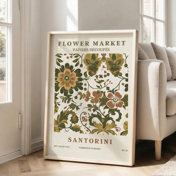 May include: Large framed botanical poster leaning against white wall next to beige sofa. Print features vintage-style floral pattern with green leaves, yellow and coral flowers on cream background. Text reads "Flower Market" and "Santorini" in green and coral lettering. Natural wood frame, bright room with windows and hardwood floors.