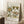 May include: Vintage-style botanical poster with decorative floral pattern in green, gold and terracotta tones, displayed in light wood frame leaning against white paneled wall. Coffee cup on wooden block and textured beige throw blanket visible nearby, creating cozy room setting with natural lighting.