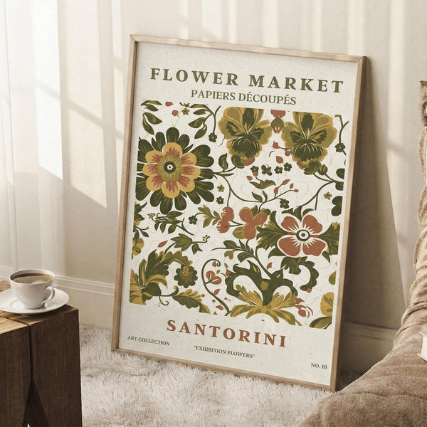May include: Vintage-style botanical poster with decorative floral pattern in green, gold and terracotta tones, displayed in light wood frame leaning against white paneled wall. Coffee cup on wooden block and textured beige throw blanket visible nearby, creating cozy room setting with natural lighting.