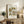 May include: Vintage-style botanical poster with colorful floral pattern in earth tones displayed in light wood frame on rustic wooden sideboard. Room setting features fern plant, beige armchair with brown leather pillow, ceramic vase, and stacked books. Warm natural lighting creates cozy home decor atmosphere.