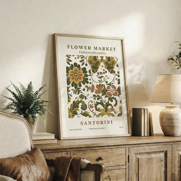 May include: Vintage-style botanical poster with colorful floral pattern in earth tones displayed in light wood frame on rustic wooden sideboard. Room setting features fern plant, beige armchair with brown leather pillow, ceramic vase, and stacked books. Warm natural lighting creates cozy home decor atmosphere.