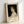 May include: Framed art print leaning against white wall showing classical portrait painting of young woman with curly dark hair in white dress against black background. Cream matting displays "PIERRE AUGUSTE exhibition" text with "Gabrielle Cot" and "Pierre Auguste's Daughter" below. Natural wood frame, soft lighting creates gentle shadows on white surface.