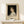 May include: Framed art print featuring a classical portrait of a young woman with dark curly hair wearing a white dress against a black background. The poster displays "PIERRE AUGUSTE exhibition" text below the portrait. Shows cream matting in natural wood frame, positioned on white wooden surface with small wrapped gift box nearby. Soft lighting creates gentle shadows on neutral wall background.