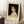 May include: Framed art print leaning against white wall on fluffy white rug next to wooden side table with coffee cup. Print shows classical portrait painting of young woman with curly hair in white dress against dark background. Cream matting with "PIERRE AUGUSTE exhibition" text below image. Soft natural lighting creates shadows on wall.