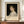 May include: Framed art print featuring a classical portrait of a young woman with curly brown hair wearing a white ruffled dress against a dark background. The poster displays "PIERRE AUGUSTE exhibition" in bold black text below the painting, with smaller text reading "Gabrielle Cot" and "Pierre Auguste's Daughter." The print sits on a wooden sideboard next to a white ceramic vase and books, displayed against cream-colored paneled walls in an elegant interior setting.