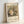 May include: Framed art print leaning against white wall showing Gustav Klimt's Baby (Cradle) painting from 1917. The artwork depicts swirling, organic forms in earth tones with intricate patterns characteristic of Klimt's style. Museum of Modern Art New York attribution visible below the image. Light wood frame with white matting, casting soft shadows on the wall.