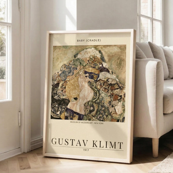 May include: Framed art print leaning against white wall next to beige sofa in bright room with windows. Print shows Gustav Klimt's colorful painting of a baby surrounded by ornate patterned fabrics and decorative elements in earth tones, blues, and golds. Cream-colored poster with black text identifying the artwork, artist name, and museum attribution. Light wood frame, approximately 2-3 feet tall, positioned on light wooden floor.