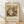 May include: Framed art print of Gustav Klimt's "Baby (Cradle)" from 1917 leaning against white wall on light wooden floor. The poster shows a swirling composition of a mother and infant rendered in Klimt's signature decorative style with ornate patterns, rich earth tones, and golden hues. Museum attribution text visible below the artwork in natural wood frame with cream matting.