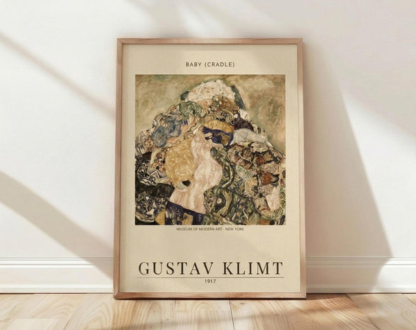 May include: Framed art print of Gustav Klimt's "Baby (Cradle)" from 1917 leaning against white wall on light wooden floor. The poster shows a swirling composition of a mother and infant rendered in Klimt's signature decorative style with ornate patterns, rich earth tones, and golden hues. Museum attribution text visible below the artwork in natural wood frame with cream matting.