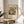 May include: Framed art print displaying Gustav Klimt's "Baby (Cradle)" painting featuring swirling, ornate patterns in earth tones surrounding pale figures. The poster has cream matting with black text identifying the artwork and artist name. Shown in thin wooden frame on round wooden side table next to beige upholstered chair with white throw pillow. Soft natural lighting creates shadows on neutral wall, with green plant leaves visible in corner.