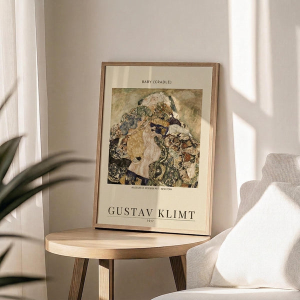 May include: Framed art print displaying Gustav Klimt's "Baby (Cradle)" painting featuring swirling, ornate patterns in earth tones surrounding pale figures. The poster has cream matting with black text identifying the artwork and artist name. Shown in thin wooden frame on round wooden side table next to beige upholstered chair with white throw pillow. Soft natural lighting creates shadows on neutral wall, with green plant leaves visible in corner.