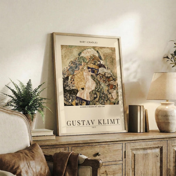 May include: Framed Gustav Klimt museum exhibition poster displaying Baby (Cradle) artwork in warm earth tones and intricate patterns, positioned on rustic wooden sideboard alongside fern plant, ceramic vase, and leather chair in cozy neutral-toned living room setting with soft natural lighting
