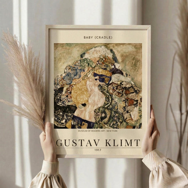 May include: Person in beige clothing holding framed art print against white curtained window, with dried pampas grass visible. The vintage-style poster features Gustav Klimt's colorful, ornate painting of a mother holding a baby, surrounded by decorative patterns in blues, golds, and earth tones. Museum exhibition format with cream matting and thin wooden frame.