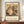 May include: Framed museum poster featuring Gustav Klimt's colorful expressionist painting of a mother and baby surrounded by swirling fabrics and patterns in earthy tones of gold, green, and brown. The poster sits on a wooden sideboard next to a white ceramic vase and books against cream walls with white molding in a modern interior setting.