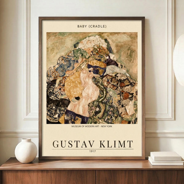 May include: Framed museum poster featuring Gustav Klimt's colorful expressionist painting of a mother and baby surrounded by swirling fabrics and patterns in earthy tones of gold, green, and brown. The poster sits on a wooden sideboard next to a white ceramic vase and books against cream walls with white molding in a modern interior setting.