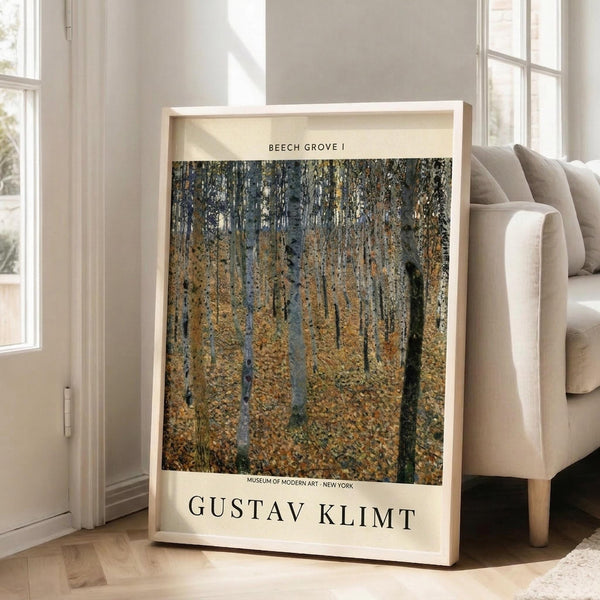May include: Framed art print of Gustav Klimt's Beech Grove I painting featuring golden autumn birch trees with white bark against yellow foliage, displayed in a light wooden frame leaning against white walls near a beige upholstered chair in a bright room with tall windows and natural lighting on light wood floors.