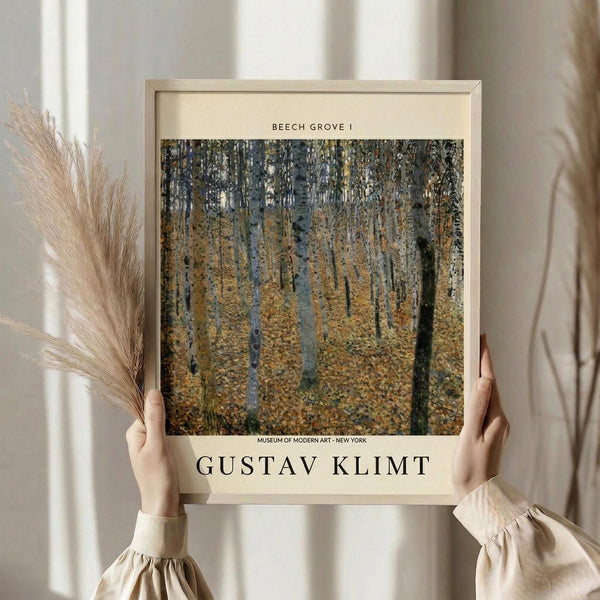 May include: Person in cream ruffled sleeves holding framed art print showing Gustav Klimt's impressionist forest scene with golden autumn leaves and silver birch tree trunks. Print has cream matting, light wood frame, with "Beech Grove I" and "Museum of Modern Art - New York" text. Dried pampas grass visible on left side against soft neutral background.