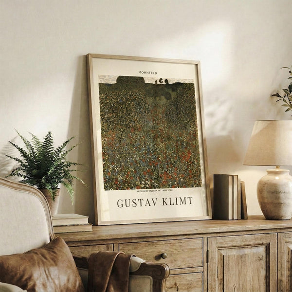 May include: Framed Gustav Klimt poster displaying a textured landscape painting with rolling green hills dotted with colorful flowers and distant trees, mounted in light wood frame. Print sits on rustic wooden sideboard alongside fern plant, ceramic vase, books, and table lamp. Beige upholstered chair with brown throw blanket visible in foreground against cream wall.