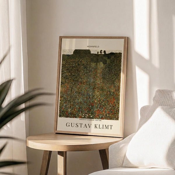 May include: Framed art print featuring Gustav Klimt's "Mohnfeld" displayed on a round wooden side table in a bright, minimalist room. The poster shows a textured landscape painting with a vibrant field of red poppies and green foliage under trees, with cream matting and thin wooden frame. Soft natural lighting creates shadows from nearby plant leaves and a beige upholstered chair.