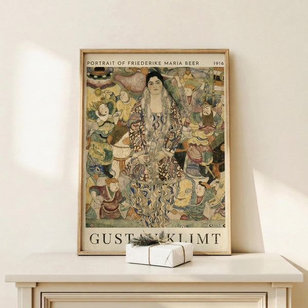 May include: Framed poster displaying Gustav Klimt's 1916 Portrait of Friederike Maria Beer, featuring a woman with long dark hair in an ornate patterned dress surrounded by colorful figures and decorative elements in Klimt's distinctive Art Nouveau style. The vintage-style print is in a light wood frame, positioned on a pale wooden shelf next to a small white gift box with evergreen sprig against a cream wall.