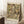 May include: Framed art print leaning against white wall corner showing Gustav Klimt's 1916 portrait featuring a woman in ornate patterned dress surrounded by colorful figures and decorative elements in his distinctive Art Nouveau style. Light wood frame, positioned on white textured rug beside wooden side table with coffee cup and saucer, beige furniture visible at edge.