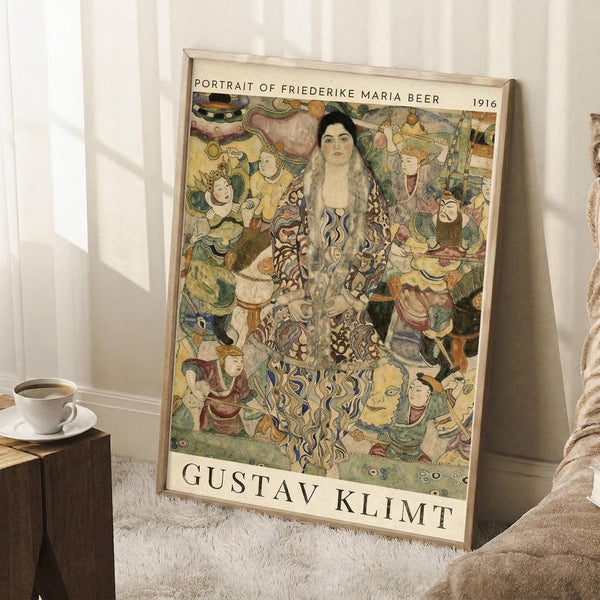 May include: Framed art print leaning against white wall corner showing Gustav Klimt's 1916 portrait featuring a woman in ornate patterned dress surrounded by colorful figures and decorative elements in his distinctive Art Nouveau style. Light wood frame, positioned on white textured rug beside wooden side table with coffee cup and saucer, beige furniture visible at edge.