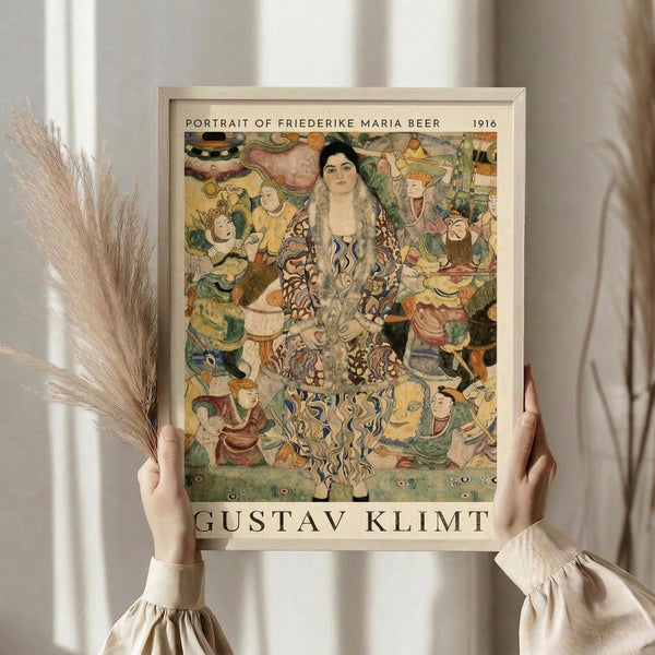 May include: Hands holding a framed art print of a colorful portrait showing a woman with long dark hair wearing an ornate patterned dress, surrounded by decorative figures and swirling motifs in vibrant yellows, greens, and earth tones. Light wooden frame against white curtained window with dried wheat stalks visible on left side.