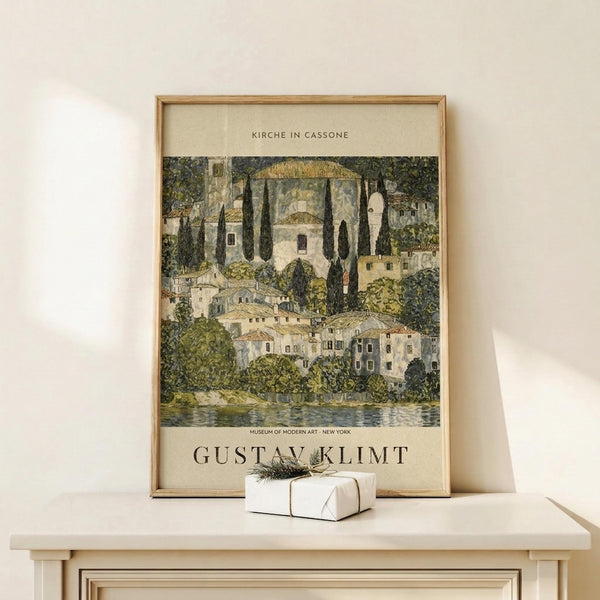 May include: Framed art print featuring a lakeside village scene with Mediterranean-style buildings, tall cypress trees, and domed structures in muted greens, whites, and earth tones. The poster shows "KIRCHE IN CASSONE" at top and "GUSTAV KLIMT" at bottom with museum attribution. Displayed on light wood surface with small white wrapped gift beside it against cream wall.