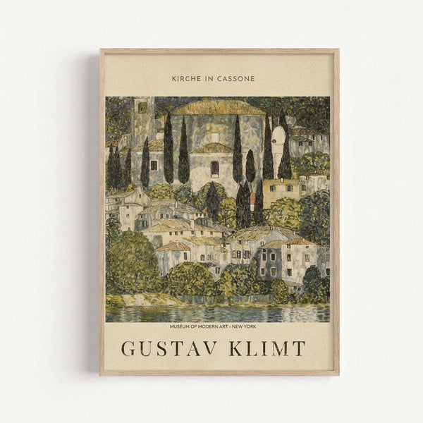 Framed art print showing a Mediterranean hillside village with white buildings, tall dark cypress trees, and golden domed structures nestled among lush green foliage beside water. The artwork features muted earth tones in an Art Nouveau style. Text at top reads "KIRCHE IN CASSONE" and bottom shows "MUSEUM OF MODERN ART - NEW YORK" above "GUSTAV KLIMT" in large letters. Light wood frame against white wall.