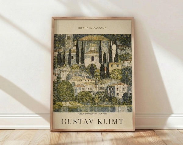 May include: Framed art print leaning against white wall on wooden floor with natural lighting. The poster features a colorful village landscape with Mediterranean-style buildings, cypress trees, a prominent church with domed architecture, and waterfront view in earthy greens, yellows, and muted tones. Light wood frame surrounds cream-colored matting with black text above and below the artwork.