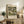 May include: Framed art print featuring a pastoral landscape with domed church buildings, cypress trees, and Mediterranean-style houses in muted greens and earth tones. The vintage-style poster displays against a cream wall above a rustic wooden sideboard with drawers and cabinet doors. Scene includes fern plant, stacked books, ceramic vase, and warm lighting creating cozy home decor atmosphere.