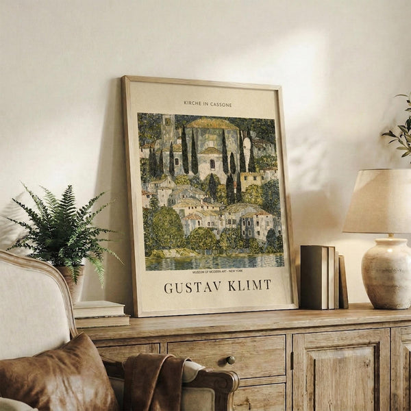 May include: Framed art print featuring a pastoral landscape with domed church buildings, cypress trees, and Mediterranean-style houses in muted greens and earth tones. The vintage-style poster displays against a cream wall above a rustic wooden sideboard with drawers and cabinet doors. Scene includes fern plant, stacked books, ceramic vase, and warm lighting creating cozy home decor atmosphere.