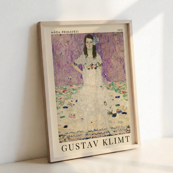 May include: Framed art print leaning against white wall showing Gustav Klimt's portrait of a young woman in white dress with floral embroidery against purple patterned background. The painting features Klimt's distinctive decorative style with colorful scattered motifs on floor and wallpaper. Light wood frame with cream matting displays the artwork title and artist name below the image.