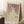 May include: Framed art print leaning against white wall on fluffy white rug next to wooden side table with coffee cup. Print shows woman in white dress with floral details against purple background, dated 1912, with Gustav Klimt text at bottom. Light wood frame, natural lighting creates soft shadows on wall.