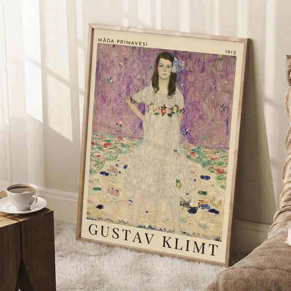 May include: Framed art print leaning against white wall on fluffy white rug next to wooden side table with coffee cup. Print shows woman in white dress with floral details against purple background, dated 1912, with Gustav Klimt text at bottom. Light wood frame, natural lighting creates soft shadows on wall.
