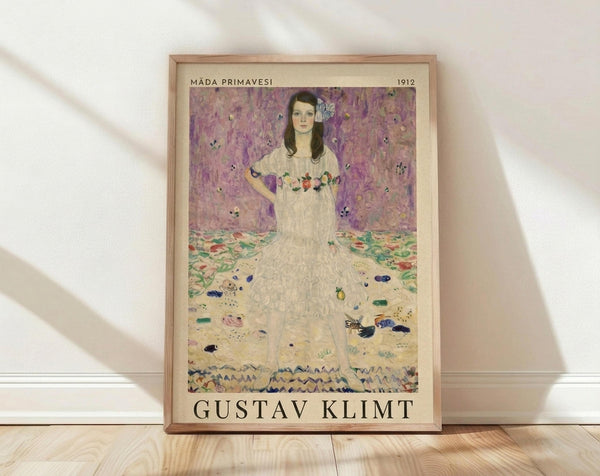 May include: Framed art print reproduction of Gustav Klimt's portrait painting featuring a young woman in an ornate white dress with floral details against a decorative purple and pink patterned background. The poster shows in a light wood frame, leaning against a white wall on wooden flooring with soft natural lighting creating diagonal shadows across the scene.