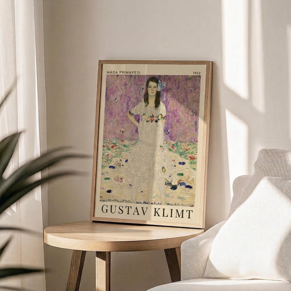 May include: Framed art print featuring a woman in a white dress with floral details against a purple and pink patterned background, displayed on a round wooden side table next to a beige upholstered chair. Natural sunlight streams across the neutral-toned room setting with a potted plant visible in the corner.
