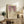 May include: Framed art print featuring a woman in white dress with floral details against purple floral wallpaper background, displayed on rustic wooden sideboard with books, ceramic vase, and fern plant. Warm neutral room setting with beige chair and soft lighting creates cozy home decor atmosphere.