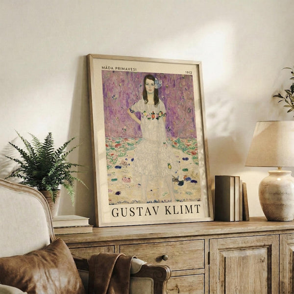 May include: Framed art print featuring a woman in white dress with floral details against purple floral wallpaper background, displayed on rustic wooden sideboard with books, ceramic vase, and fern plant. Warm neutral room setting with beige chair and soft lighting creates cozy home decor atmosphere.